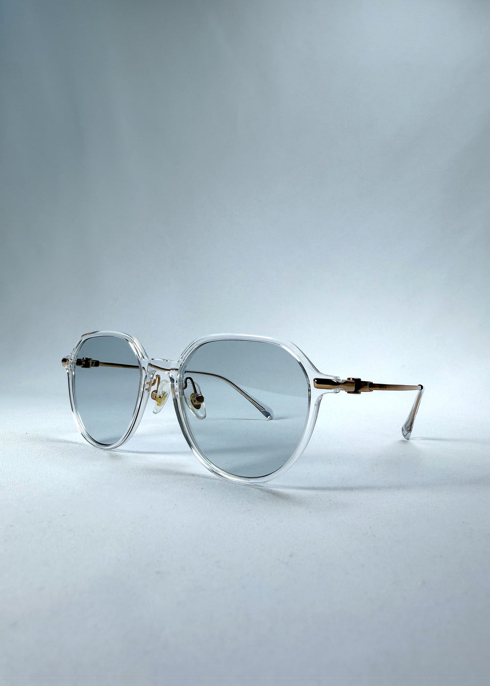 Three-quarter front view of AZE Vireo Arc sunglasses in Crystal Static, featuring a crystal-clear frame, pale blue lenses, and gold-tone accents, showing the frame shape and temple profile.