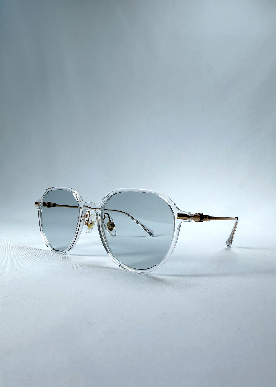 Three-quarter front view of AZE Vireo Arc sunglasses in Crystal Static, featuring a crystal-clear frame, pale blue lenses, and gold-tone accents, showing the frame shape and temple profile.