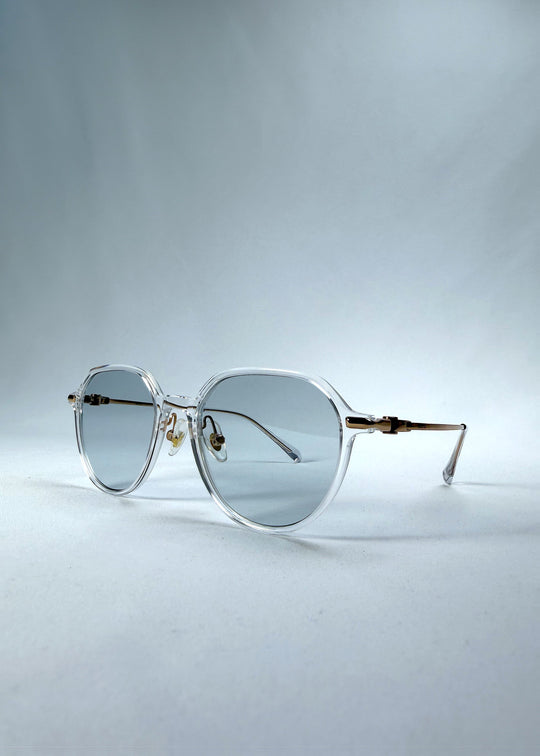 Three-quarter front view of AZE Vireo Arc sunglasses in Crystal Static, featuring a crystal-clear frame, pale blue lenses, and gold-tone accents, showing the frame shape and temple profile. 