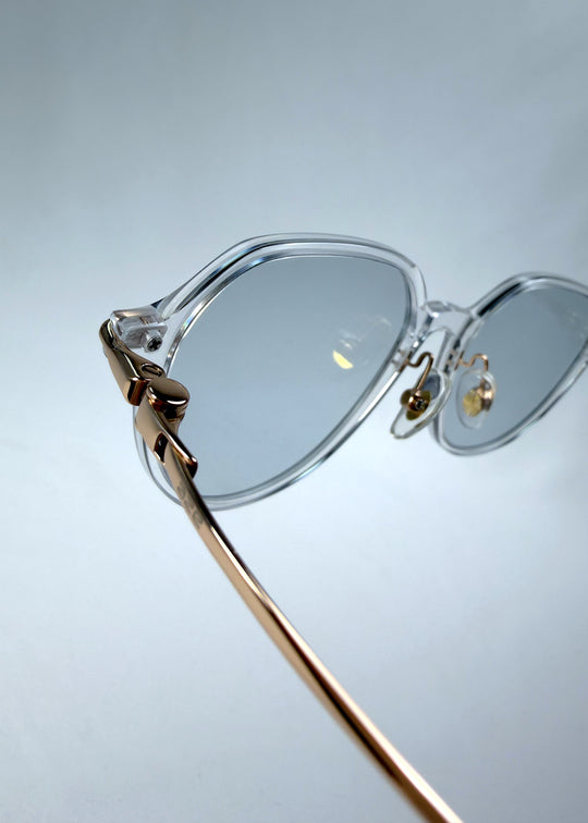 Close-up hinge detail view of AZE Vireo Arc sunglasses in Crystal Static, featuring a crystal-clear frame, pale blue lenses, and gold-tone accents, showing the hinge construction, lens edge, and temple connection.