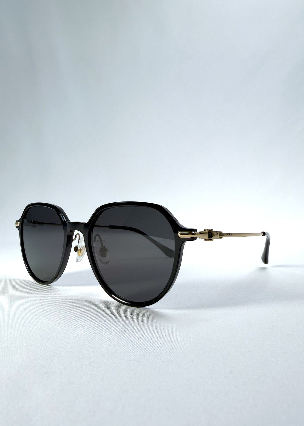 Three-quarter front view of AZE Vireo Arc sunglasses in Duskgold, featuring a dark softly geometric frame, gold-tone accents, and smoky gray lenses, showing the frame shape and temple profile.
