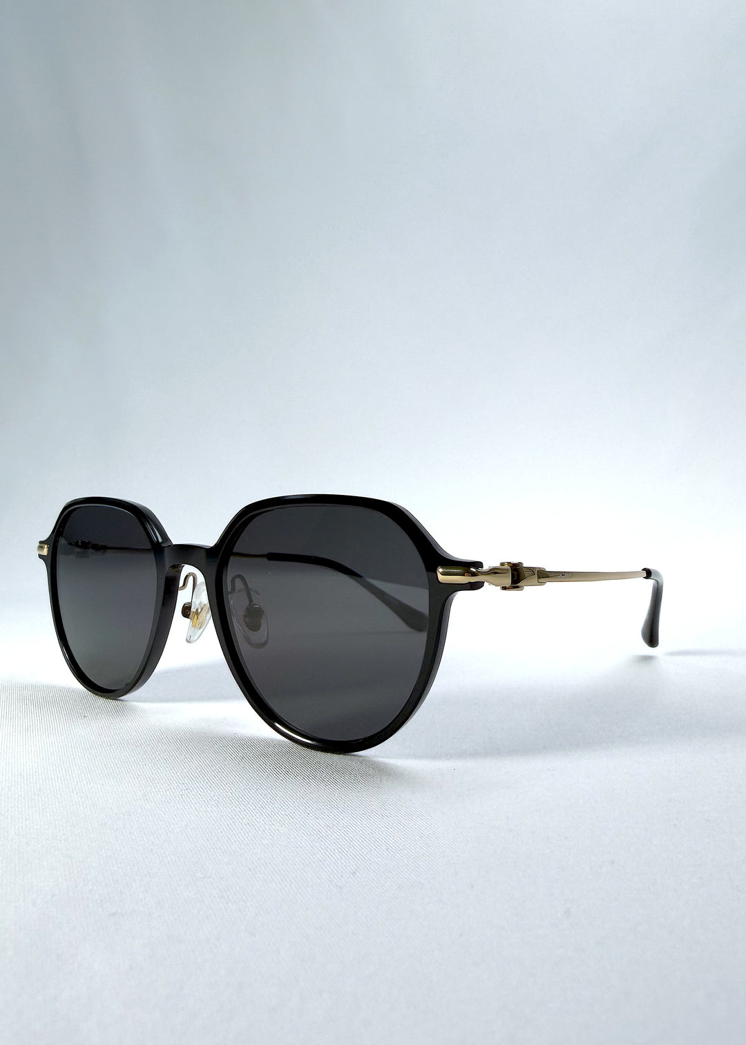Three-quarter front view of AZE Vireo Arc sunglasses in Duskgold, featuring a dark softly geometric frame, gold-tone accents, and smoky gray lenses, showing the frame shape and temple profile.