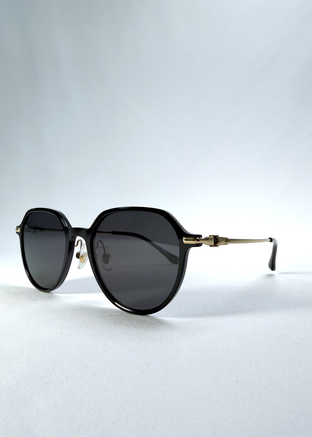 Three-quarter front view of AZE Vireo Arc sunglasses in Duskgold, featuring a dark softly geometric frame, gold-tone accents, and smoky gray lenses, showing the frame shape and temple profile. #color_duskgold