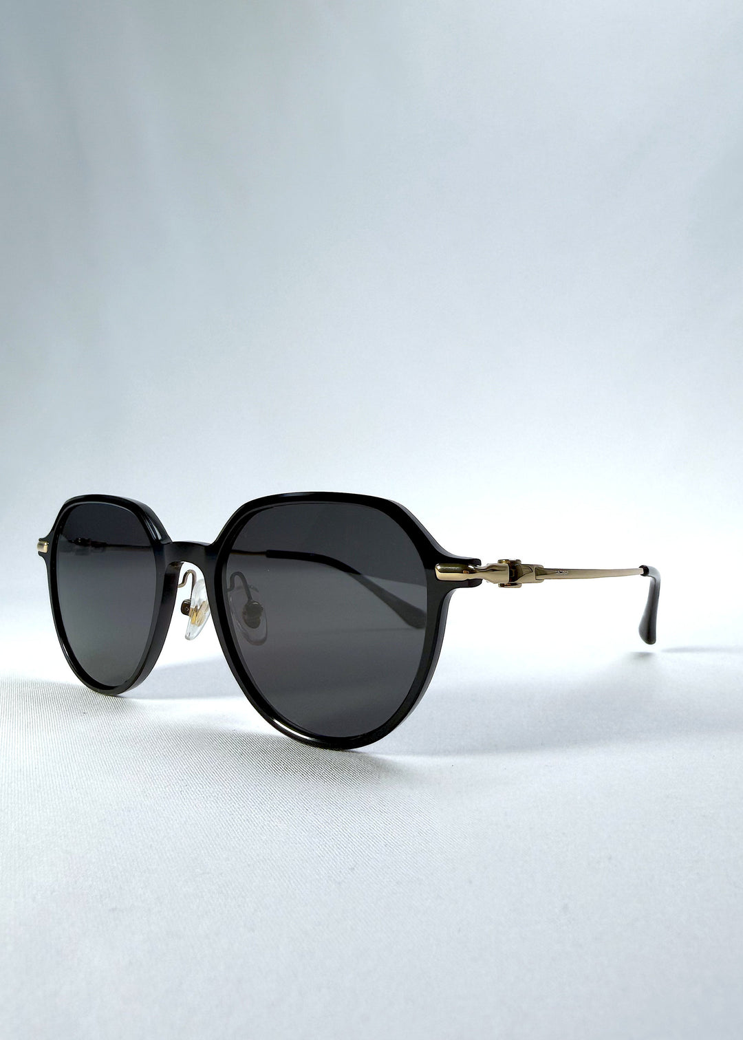 Three-quarter front view of AZE Vireo Arc sunglasses in Duskgold, featuring a dark softly geometric frame, gold-tone accents, and smoky gray lenses, showing the frame shape and temple profile. 