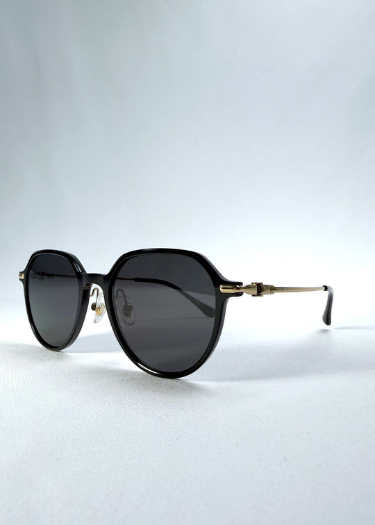 Three-quarter front view of AZE Vireo Arc sunglasses in Duskgold, featuring a dark softly geometric frame, gold-tone accents, and smoky gray lenses, showing the frame shape and temple profile. 