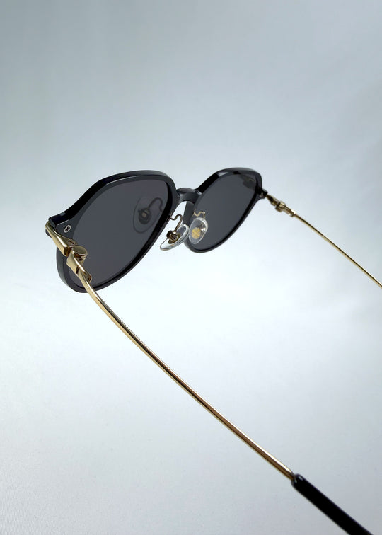 Close-up hinge detail view of AZE Vireo Arc sunglasses in Duskgold, featuring a dark softly geometric frame, gold-tone accents, and smoky gray lenses, showing the hinge construction, lens edge, and temple connection.