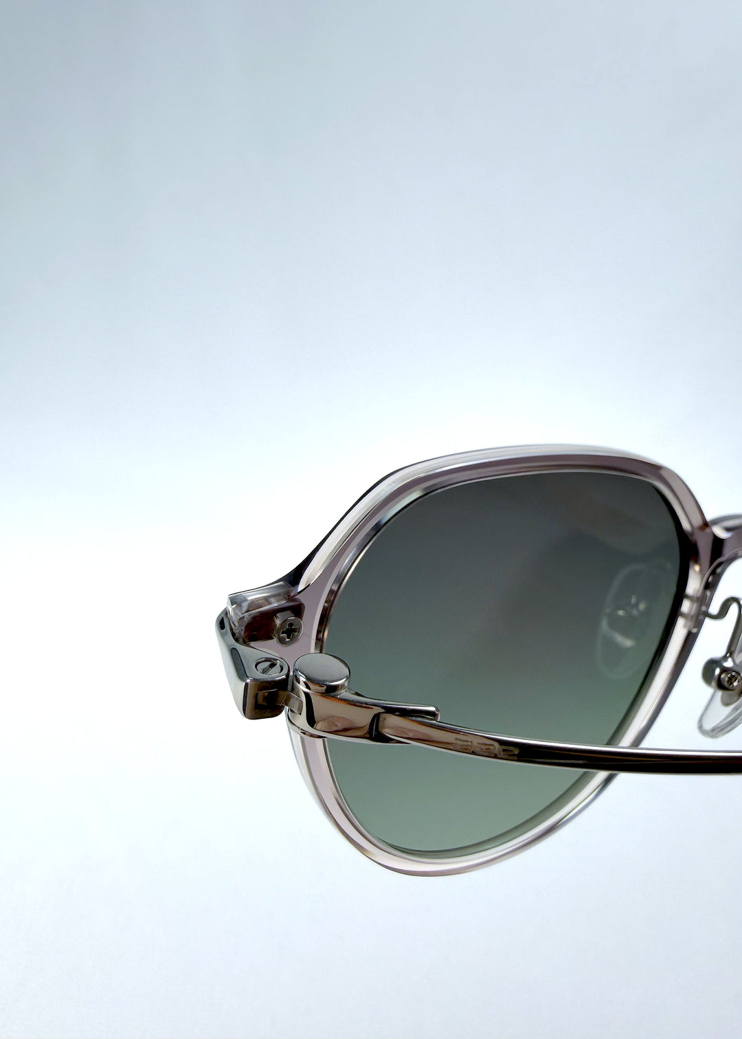 Close-up hinge detail view of AZE Vireo Arc sunglasses in Verdant Halo, featuring a translucent smoky mauve frame and green gradient lenses, showing the hinge construction, lens edge, and temple connection. 
